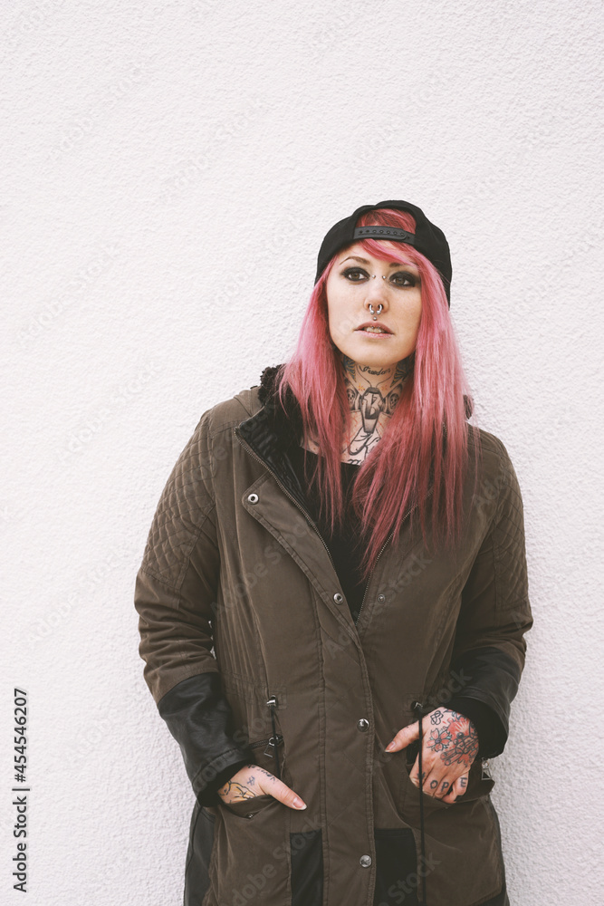 young woman with pink hair piercings and tattoos leaning against wall