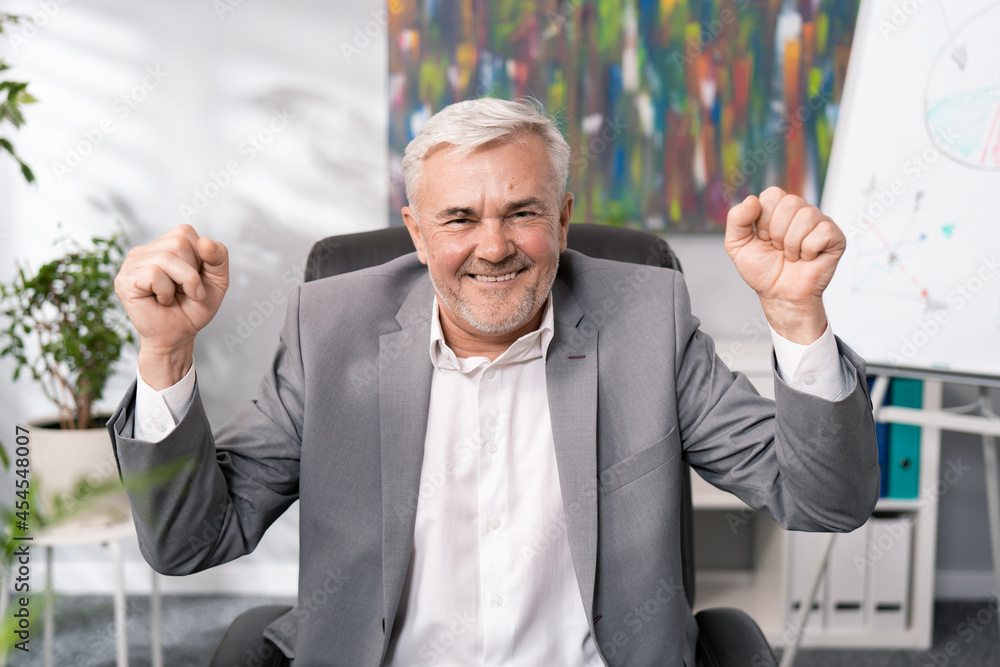 A cheerful smiling businessmen in suit with gray hair, elderly, sitting in office chair, raises hands in the air clenched in fists, the head of company won tender, signing contract, victory, happiness A cheerful smiling businessmen in suit with gray hair, elderly, sitting in office chair, raises hands in the air clenched in fists, the head of company won tender, signing contract, victory, happiness