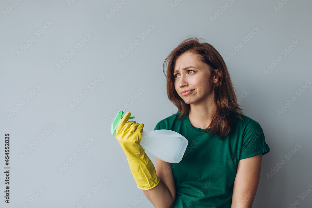 Dissatisfied young woman holding spray detergent while wearing yellow rubber gloves for hands protection during cleaning isolated over grey background copyspace Dissatisfied young woman holding spray detergent while wearing yellow rubber gloves for hands protection during cleaning isolated over grey background copyspace