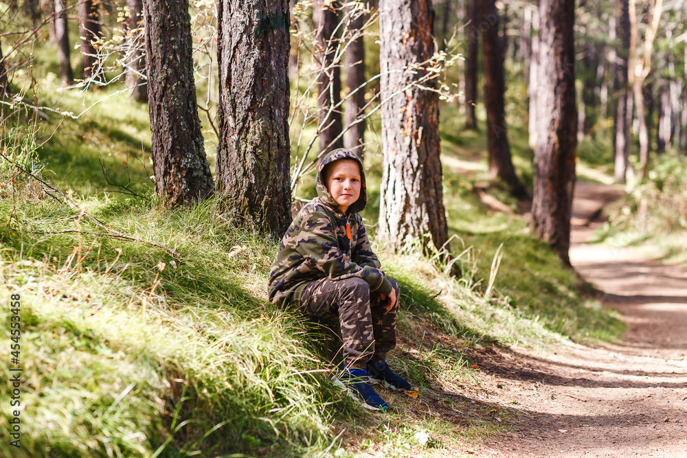 a nine-year-old boy in camouflage suit sits on a path in the woods. autumn hike