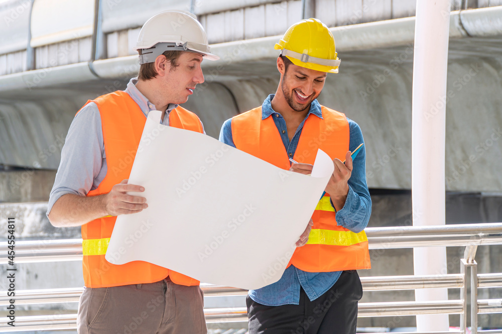 Two engineer or architect with safety helmet planning about building with blueprint. Professional technician man pointing finger while his colleagues looking at construction site.Industry work concept Two engineer or architect with safety helmet planning about building with blueprint. Professional technician man pointing finger while his colleagues looking at construction site.Industry work concept
