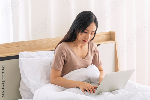 Lifestyle technology network job or marketing or e-commerce and education concept. Freelancer young woman using laptop working online while sitting alone on white bed in morning at home.