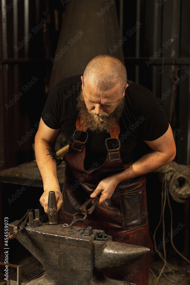 Bearded man, blacksmith manually forging the molten metal on the anvil in smithy with spark fireworks. Concept of labor, retro professions, family business Bearded man, blacksmith manually forging the molten metal on the anvil in smithy with spark fireworks. Concept of labor, retro professions, family business