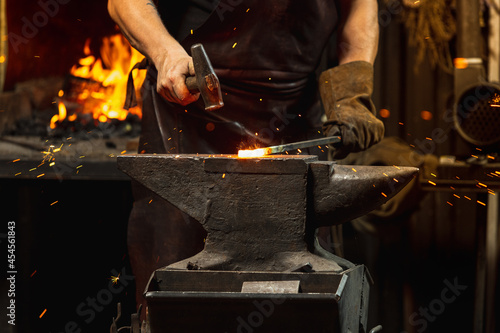 Fényképezés Bearded man, blacksmith manually forging the molten metal on the anvil in smithy with spark fireworks