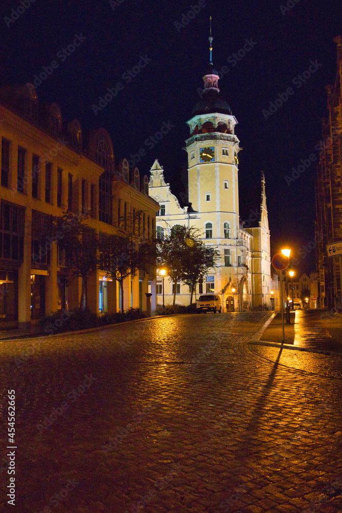 Fototapeta premium Perspektivische Nachtaufnahme vom Marktplatz Köthen mit historischer Häuserzeile und beleuchtetem Rathausturm über glänzendem Kopfsteinpflaster, Sachsen-Anhalt, Deutschland