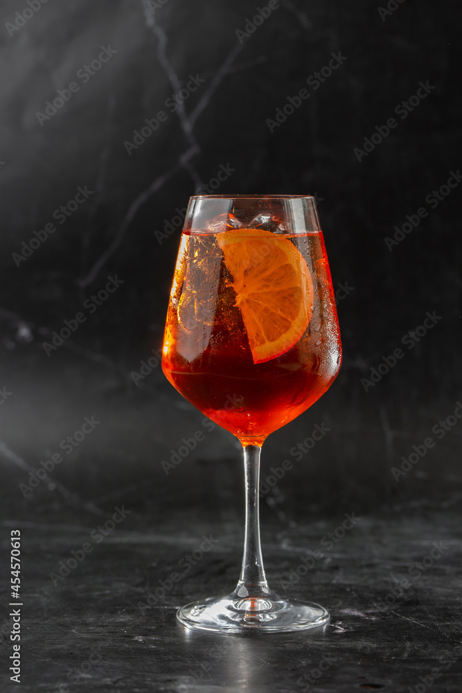 Glass of ice cold Aperol spritz cocktail served in a wine glass, decorated with slices of orange and basil leaf, placed on a black background
