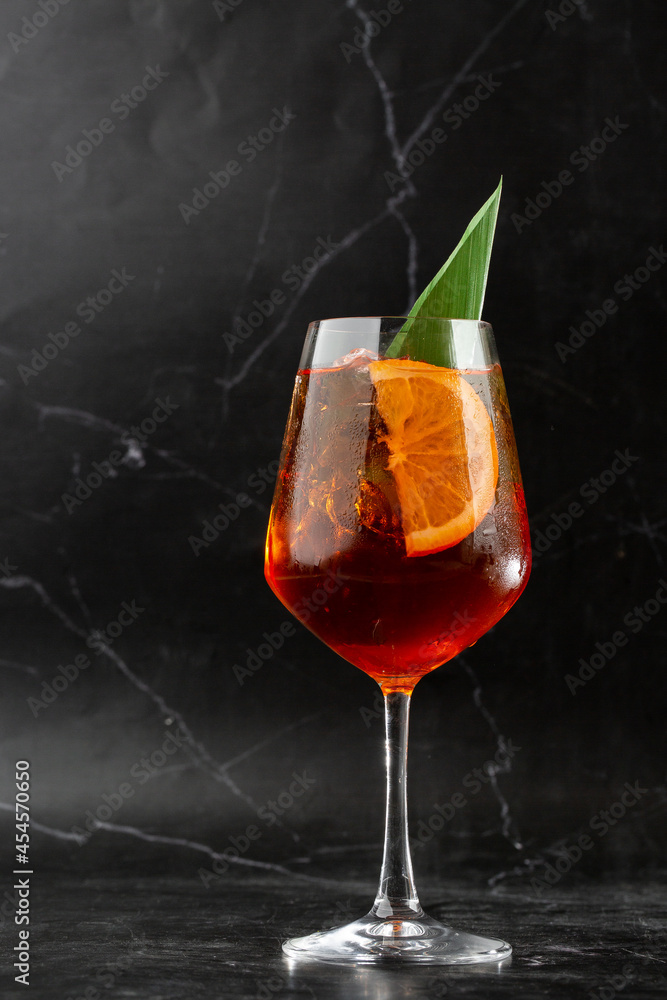 Glass of ice cold Aperol spritz cocktail served in a wine glass, decorated with slices of orange and basil leaf, placed on a black background