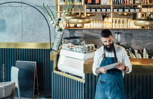Happy Barista Using Digital Tablet in a Cafe.
 
Cheerful smiling waiter with a beard leaning on the bar counter and reading online order or watching something on a digital tablet while working.
