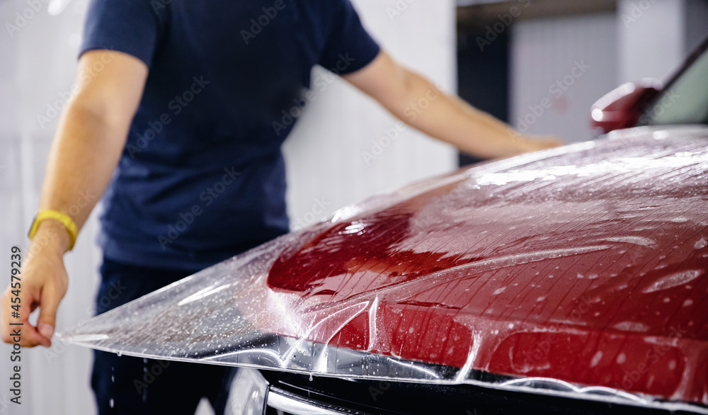 Process of pasting hood of red car with protective vinyl film from gravel chips and scratches. Transparent protection for paint