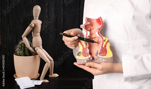 Doctor pointing at model of rectum with hemorrhoid, closeup. Wooden human figure, cactus and suppositories on black table photo