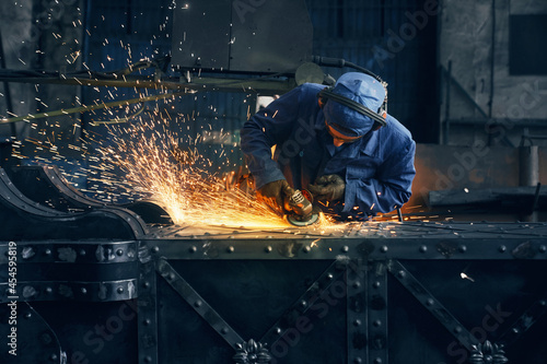 Fototapete Competent industrial worker dressed in protective clothes, glasses and gloves grinding metal construction using polishing machine