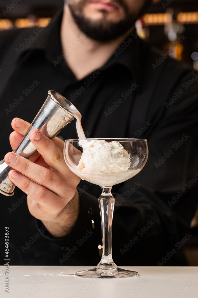 male bartender pouring to the measuring glass cup with ice cream an alcoholic drink from steel jigger on the bar counter on the blurred background