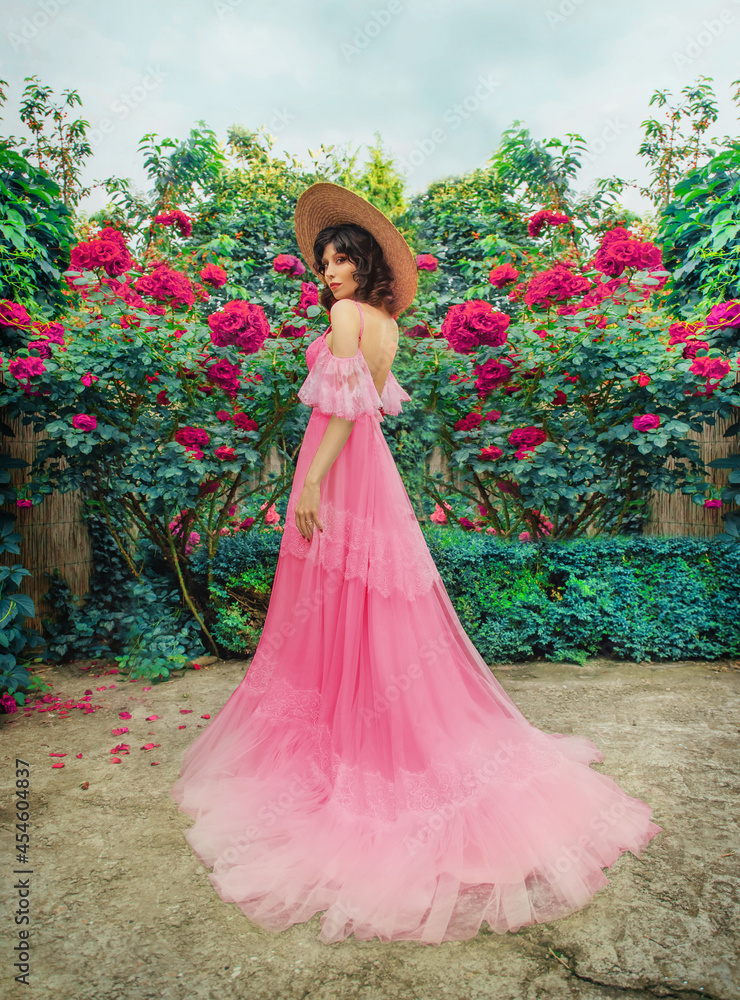 fantasy woman in pink vintage style boho dress, stands. Fashion model girl back rear view. Straw hat wide brim. Summer nature green grass trees bushes blooming rose flowers. elegant long evening gown.