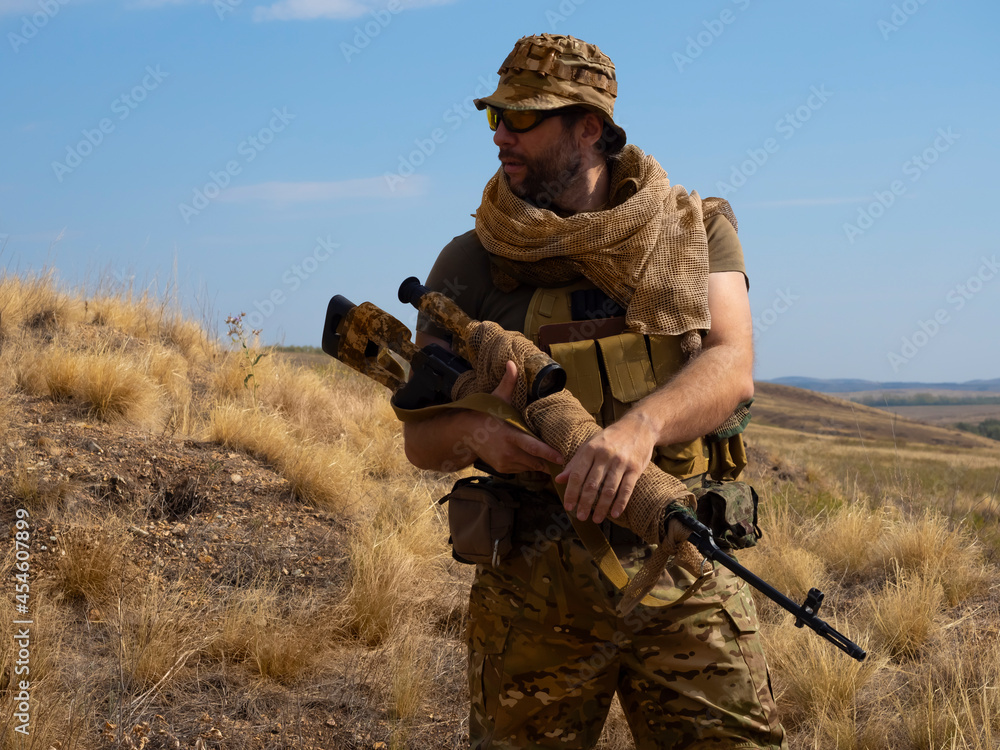 Fototapeta premium Mercenary sniper in camouflage clothes under the scorching sun. He stands with a rifle and surveys the area.