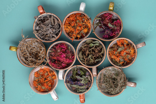 Assortment of dried relaxing tea herbs in colourful cups on blue background close up. Calendula, mint, anise hyssop, monarda didyma, wormwood, sage leaves.
