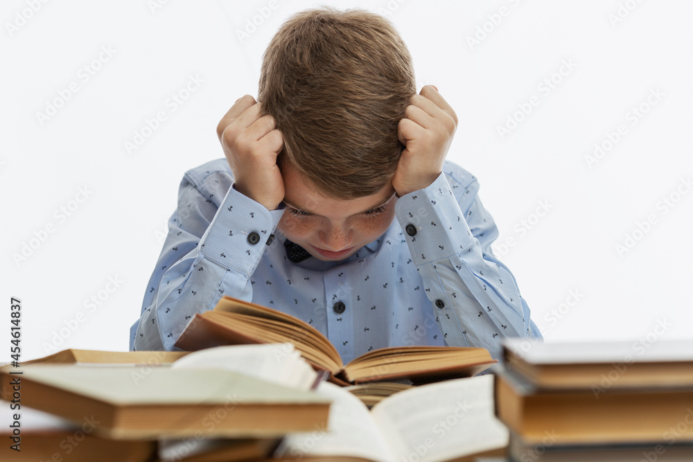 Sad boy sits at a table with many books. A child of 9-10 years old in a blue shirt clasped his head in his hands. White background. Sad boy sits at a table with many books. A child of 9-10 years old in a blue shirt clasped his head in his hands. White background.
