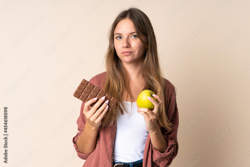 Young Lithuanian woman isolated on beige background taking a chocolate tablet in one hand and an apple in the other Young Lithuanian woman isolated on beige background taking a chocolate tablet in one hand and an apple in the other
