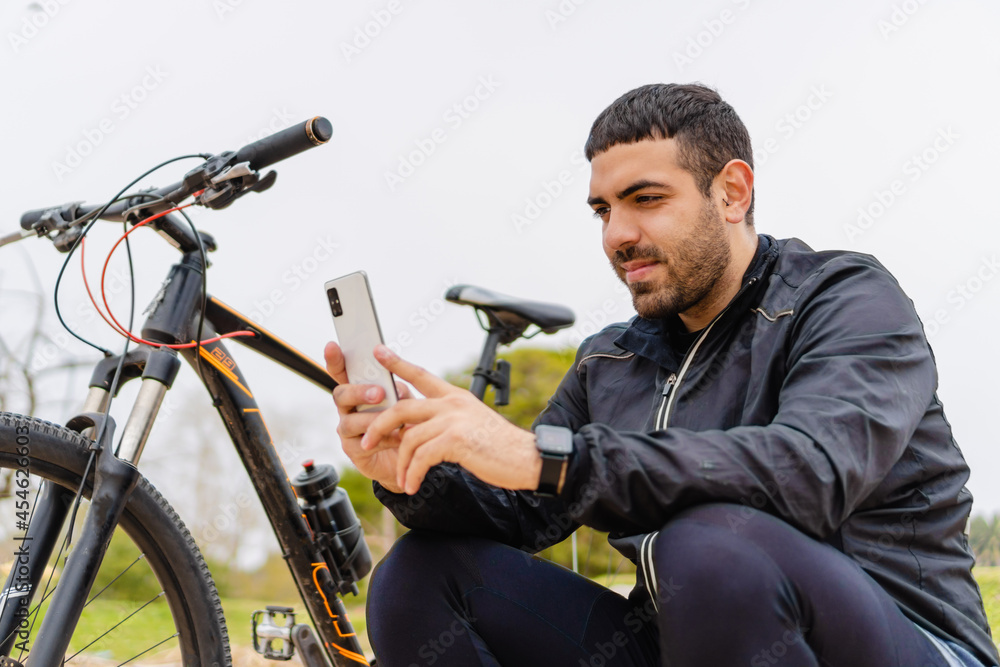 young latin hispanic man wearing a smartwatch while taking a selfie next to his bike in a park. young latin hispanic man wearing a smartwatch while taking a selfie next to his bike in a park.