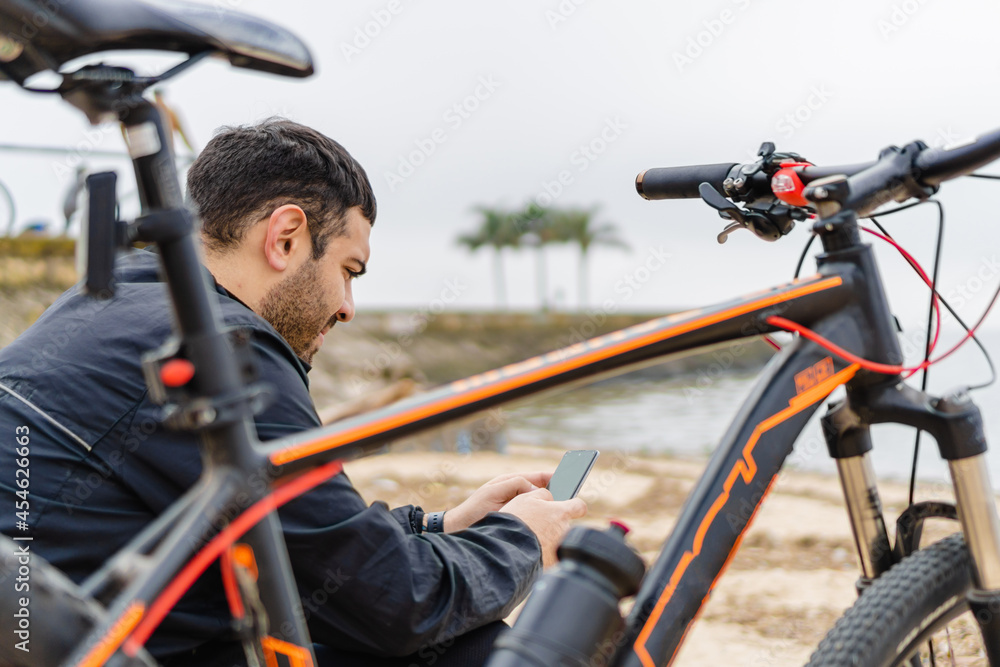 Naklejka premium young latin hispanic man checking his cell phone in a park by the river next to his bicycle.