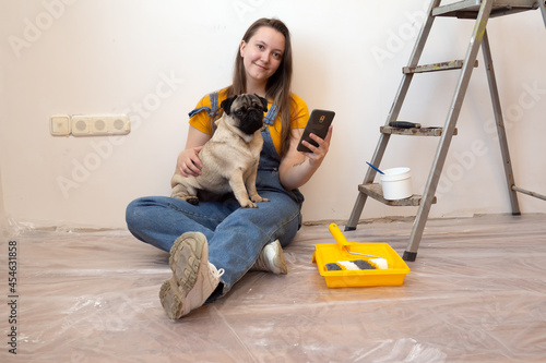 Happy woman blogger sitting on floor with pet dog in her new house during renovation, construction tools and ladder on the background. Independent single female life with pet 