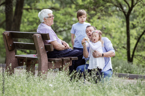 Three generation family blowing dandelion seeds in summer park: grandfather sitting on bench, father, and grandchildren