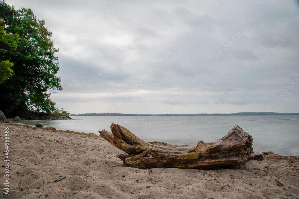 A lone log rests on a sandy beach in the 1000 Islands area of the St. Lawrence River on a gloomy and overcast day.