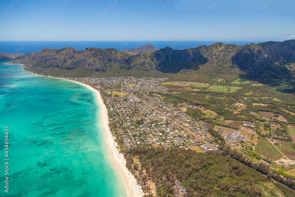 Fototapeta premium Aerial view of the coastline of the island of Oahu in Hawaii, USA. Turquoise blue water with visible reefs on left of frame, housing with green and brown mountains on the right side of the frame.