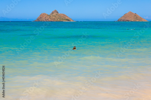 Landscape view of the twin brown Mokolua islands "Mokes" off the coast of Lanikai Beach in Oahu, Hawaii, USA. White sand beach, turquoise water, copy space in clear blue sky. One young woman in water.