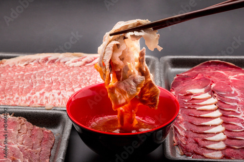 Chopsticks are picking up cooked meat and dipping in the dipping sauce. Sliced raw beef place in a row on a black plastic tray for cooking, fresh meat for grilling, yakiniku, sukiyaki or shabu. photo