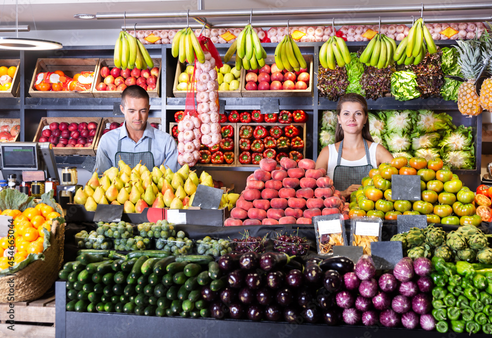Young man and woman sellers in aprons standing near counter with the fresh vegetables and fruits at market