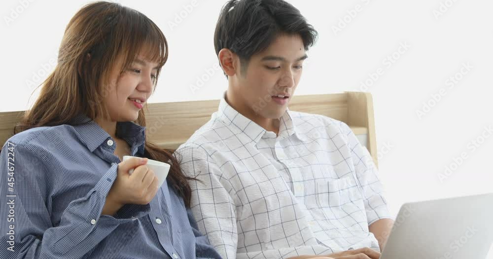 Young and cute lovers, Asian man and woman sitting on bed and using laptop together while female holding cup of coffee. Young and cute lovers, Asian man and woman sitting on bed and using laptop together while female holding cup of coffee.