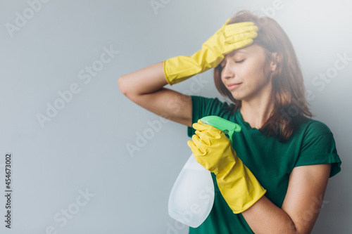 Dissatisfied young woman holding spray detergent while wearing yellow rubber gloves for hands protection during cleaning isolated over grey background copyspace 