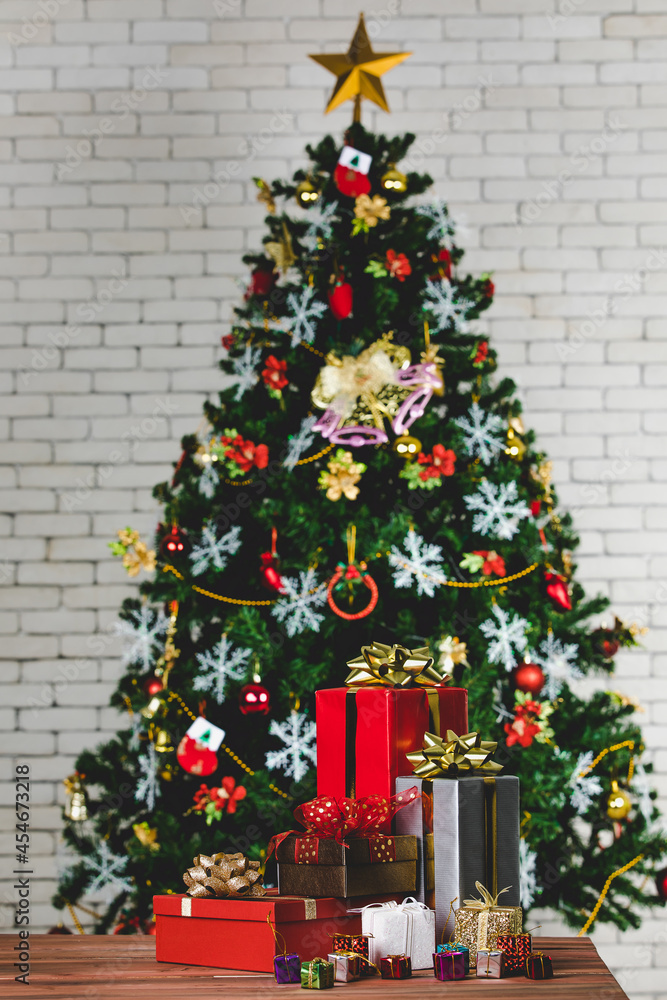 Vertical studio shot of colorful small and big present gift boxes with shiny ribbon bow tie placed on wooden table in front fully decorative beautiful Christmas eve pine tree in blurred background