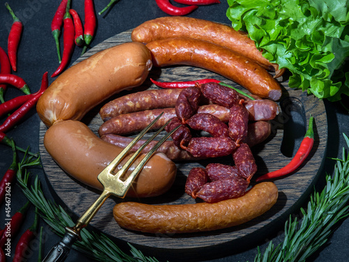 four types of pork fat smoked sausages (salami) are lying on a wooden cutting board, next to red fresh chili peppers and paprika with lettuce, a three-pronged fork is on top