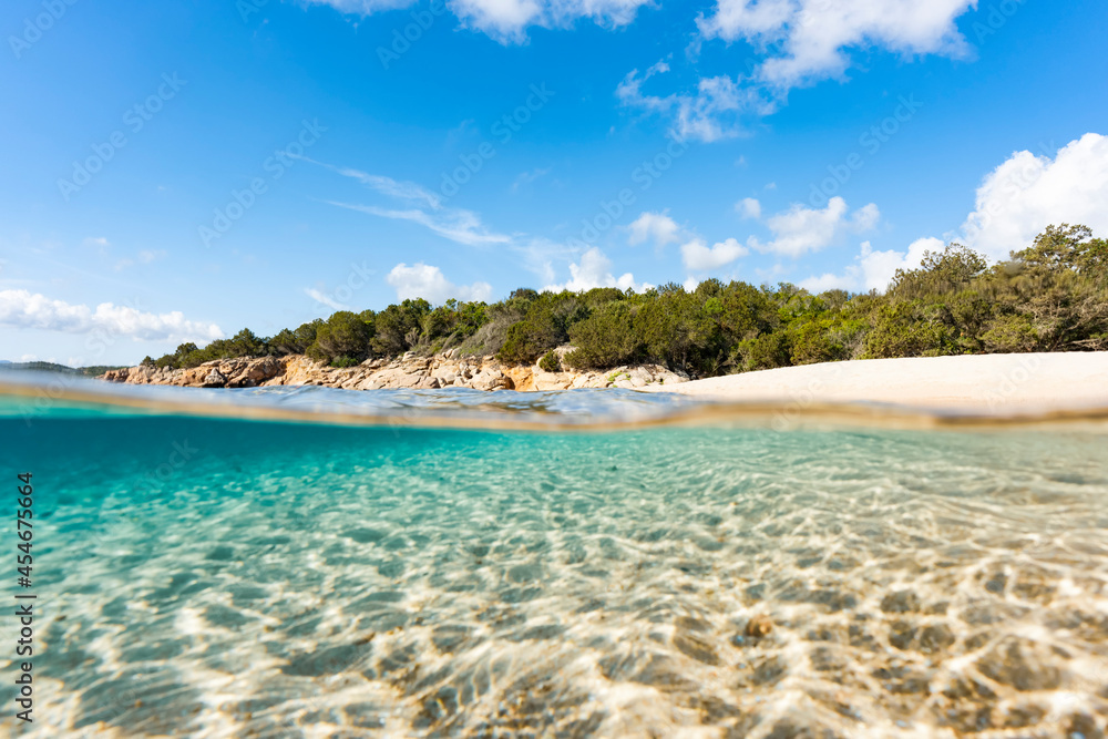 (Selective focus) Split-shot, over-under shot. Half underwater half sky with turquoise sea and a white sand beach with green vegetation. Liscia Ruja, Sardinia, Italy.