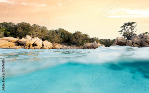 (Selective focus) Split-shot, over-under shot. Half underwater half sky with turquoise sea and a rocky coastline illuminated at sunset. Capriccioli beach, Sardinia, Italy.