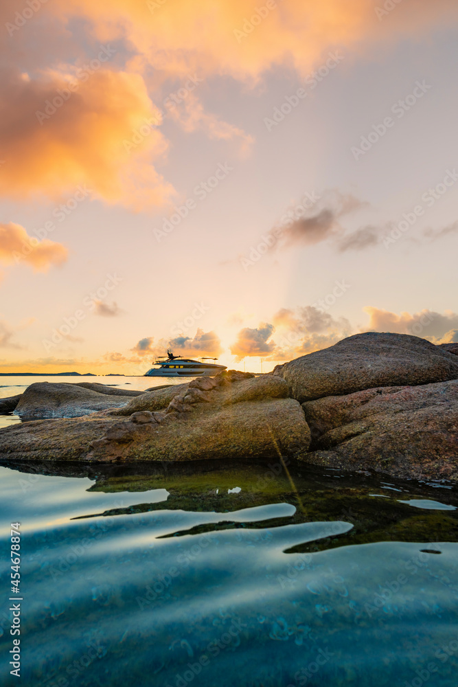 (Selective focus) Stunning view of some granite rocks in the foreground and a luxury yacht in the distance sailing on a calm sea during a dramatic sunrise. Sardinia, Italy.
