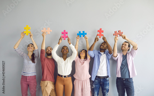 Group of happy smiling multiracial young people holding colorful puzzle pieces over their heads. Creative people stand on a light wall background. Concept of teamwork and cooperation.