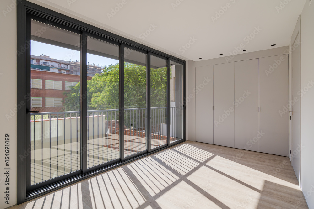 Obraz premium Sunlight illuminates the room, creating shadows on the floor. New room in a refurbished apartment with wooden floor, wardrobe and big windows.