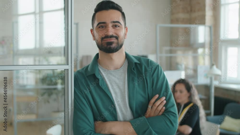Waist up portrait of positive middle eastern businessman in casualwear leaning on glass wall in office, holding arms crossed and smiling at camera Waist up portrait of positive middle eastern businessman in casualwear leaning on glass wall in office, holding arms crossed and smiling at camera