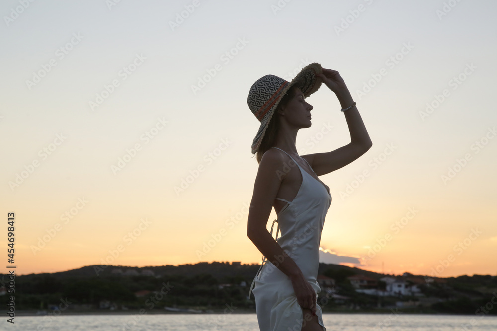 Fototapeta premium Young woman in long white dress and straw hat on the beach during sunset. Stylish summer vacation fashion.