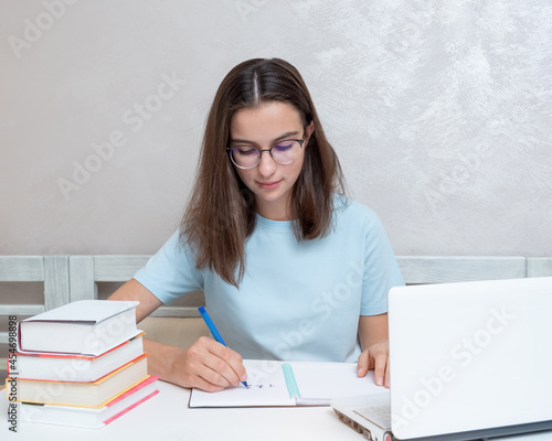 A smiling girl student sitting at a table with a laptop writes a task in a notebook. Girl student studies at home.