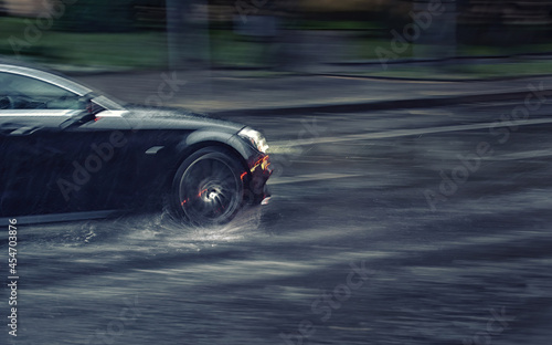 Car driving fast through puddle at heavy rain, dangers of aquaplaning, water splashing over the car. Car driving at heavy rain in evening. Dangerous driving conditions. MOTION BLUR