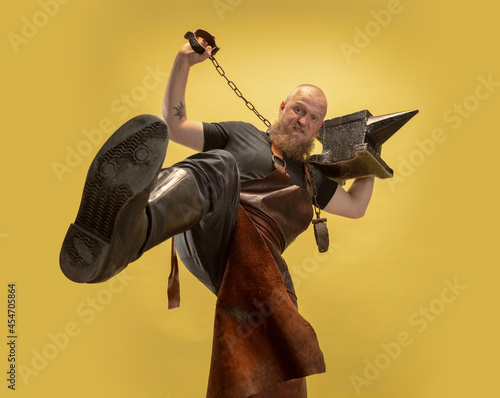 Fotografija Bottom view of muscular bearded bald man, blacksmith in leather apron or uniform isolated on yellow studio background
