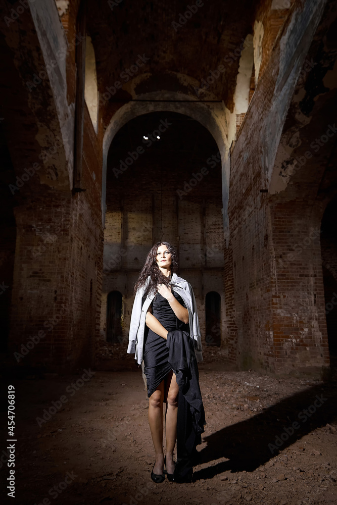 A brunette girl with long hair in an evening dress in a vintage Gothic castle. A woman who looks like an evil witch or sorceress in an abandoned old church. Female Model posing in Halloween