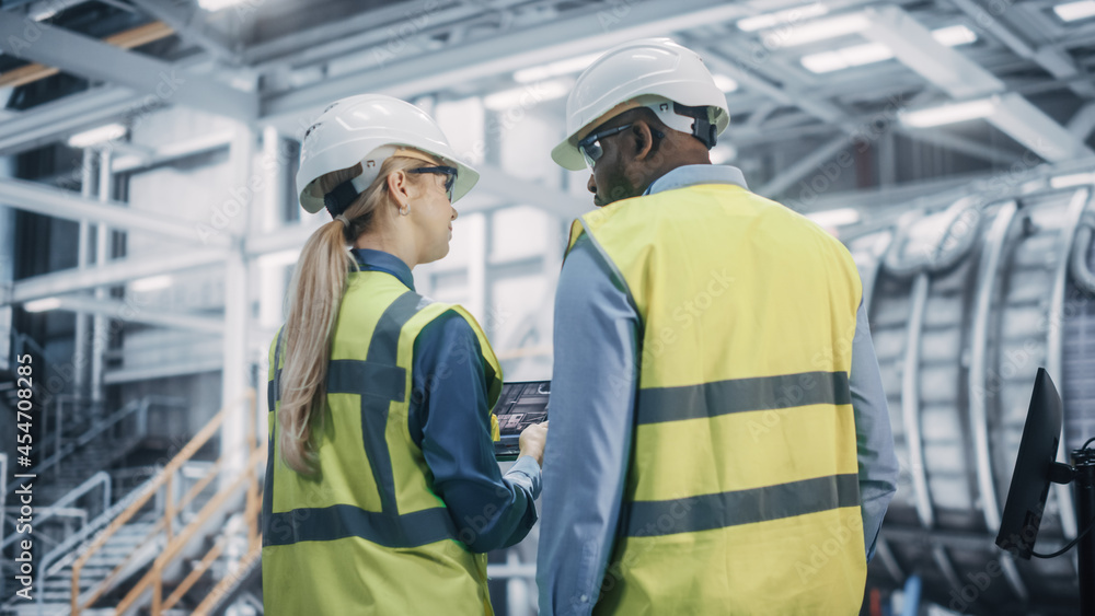 Two Diverse Professional Heavy Industry Engineers Wearing Safety Uniform and Hard Hats Working on Laptop Computer. African American Technician and Female Worker Talking on a Meeting in a Factory. Two Diverse Professional Heavy Industry Engineers Wearing Safety Uniform and Hard Hats Working on Laptop Computer. African American Technician and Female Worker Talking on a Meeting in a Factory.
