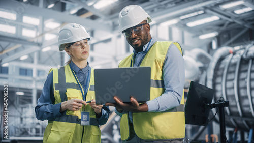 Two Diverse Professional Heavy Industry Engineers Wearing Safety Uniform and Hard Hats Working on Laptop Computer. African American Technician and Female Worker Talking on a Meeting in a Factory.