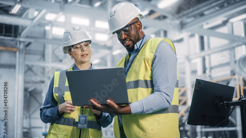 Team of Diverse Professional Heavy Industry Engineers Wearing Safety Uniform and Hard Hats Working on Laptop Computer. African American Technician and Female Worker Talking on a Meeting in a Factory.