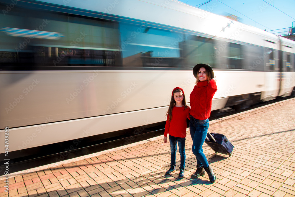 Cute sympathetic beautiful smiling young mother with daughter and suitcase on train background on railway station platform