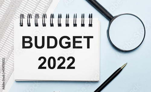 On a light blue background, reports, a magnifying glass, a pen and a notebook with the text BUDGET 2022. Business concept. Flat lay.